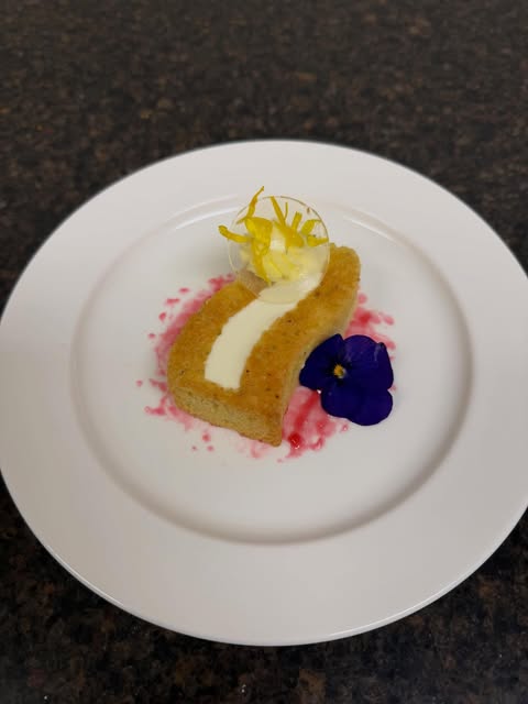 Curved golden dessert with a pale cream centre, purple flower and fine yellow garnish on a white plate.