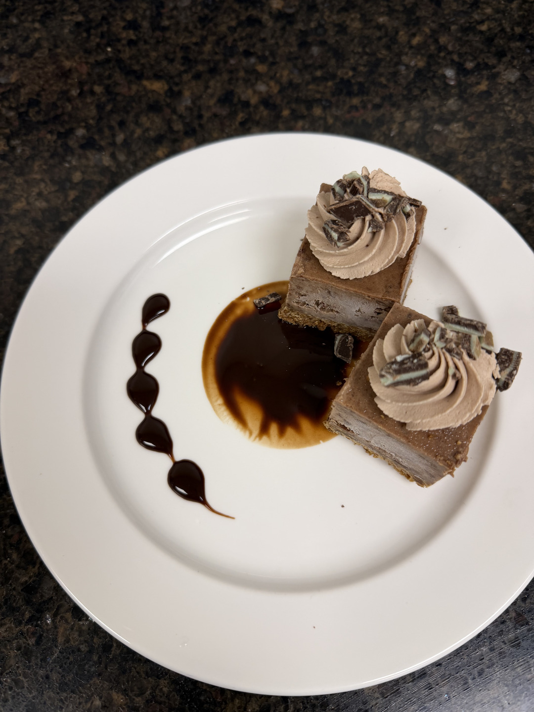 Two square chocolate mint cheesecake portions on a white plate, finished with piped rosettes, chocolate pieces and dark sauce.
